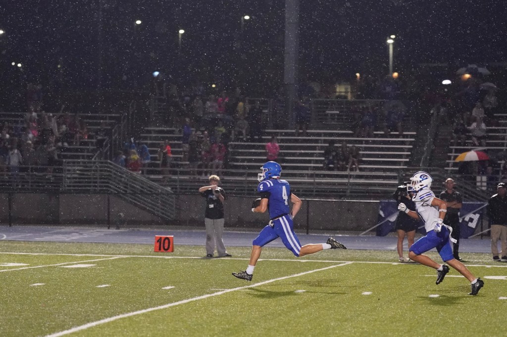 A high school football player in a blue jersey runs on the field during a rainstorm, with spectators in the background. Another player in white is pursuing him.
