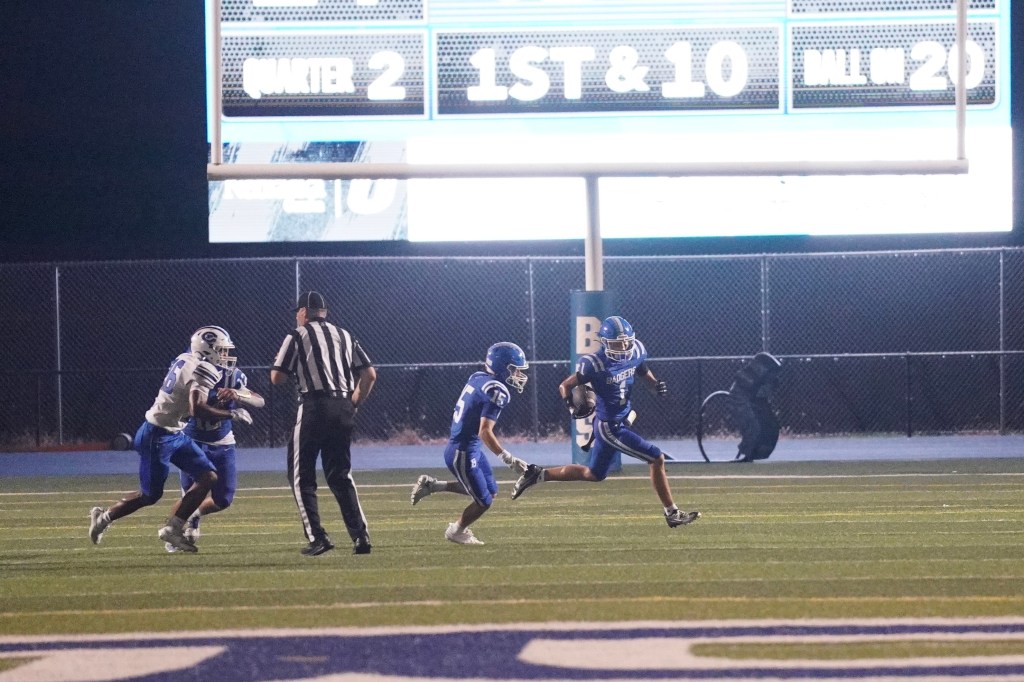 A football game under the lights with players in blue and white uniforms. Two blue players are in pursuit of a player in a white uniform, while a referee observes the play. The scoreboard displays the down and quarter.