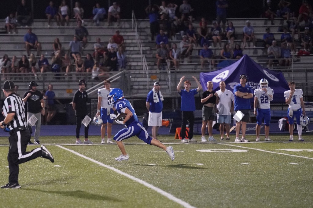 A football player in a blue uniform runs with the ball during a night game, with cheering spectators in the background.