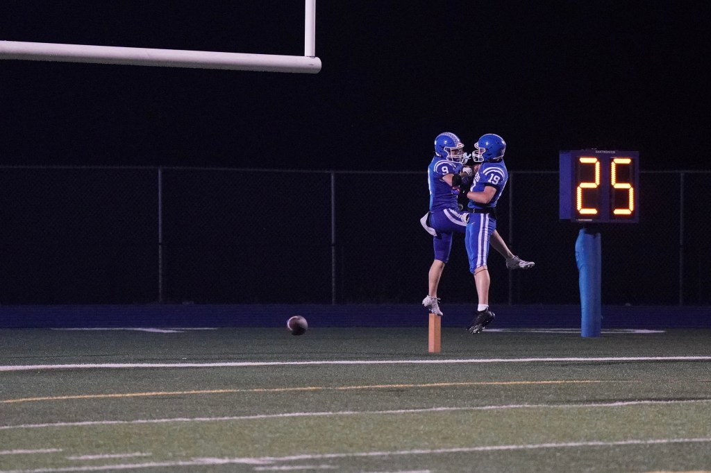Two football players in blue uniforms celebrate a touchdown by jumping in the air, with one lifting the other. A digital scoreboard displays the number 25 in the background.