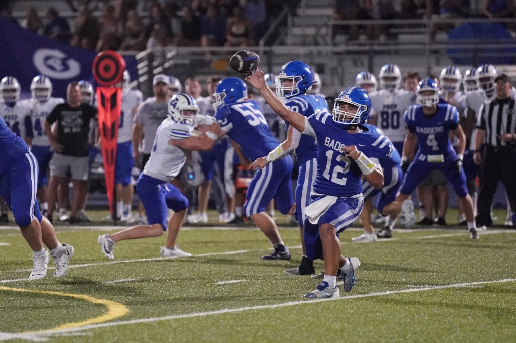 A high school football player in a blue jersey with the number 12 throws a football while being pursued by defenders on the field.