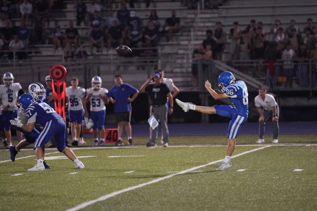 A football player in a blue uniform kicks the ball during a game, while teammates and opponents are positioned on the field.