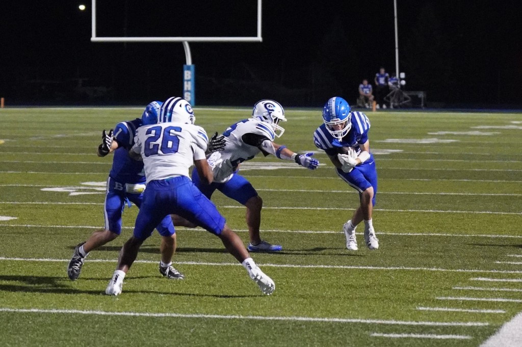 High school football action on a night game, featuring players in blue and white uniforms engaged in a play on the field.