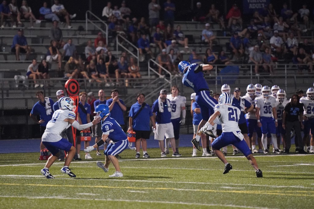 A high school football player in a blue jersey jumps to catch a pass while another player in a white jersey attempts to tackle him. Spectators can be seen in the background, with coaches and teammates watching from the sidelines.