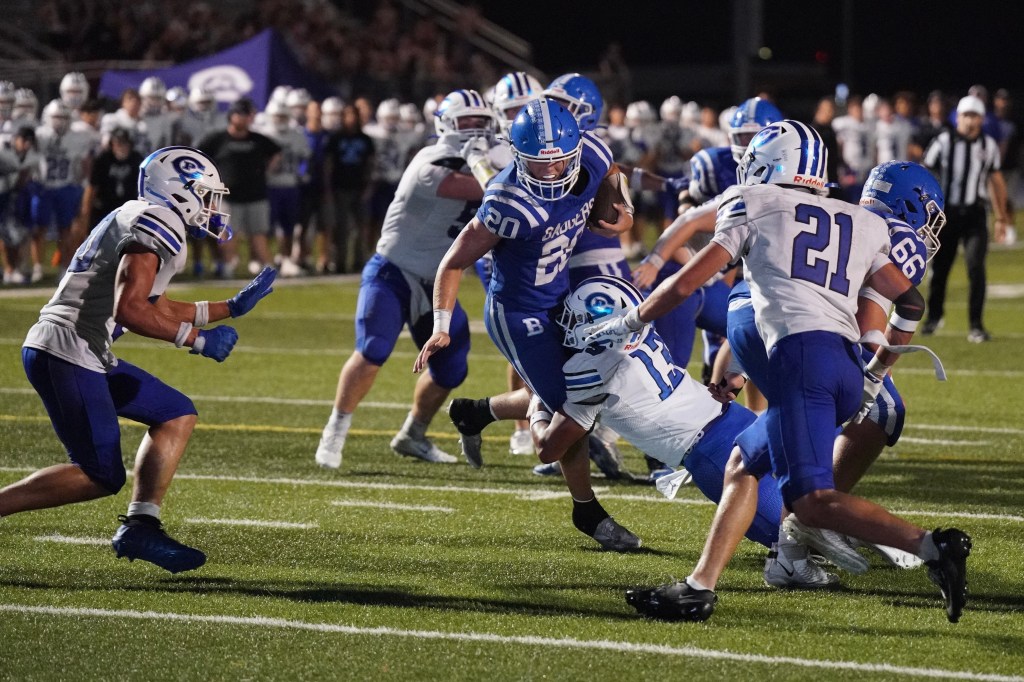 A high school football game in action, featuring players in blue and white uniforms. A player wearing number 20 is running with the ball while being tackled by opponents, with a crowd visible in the background.