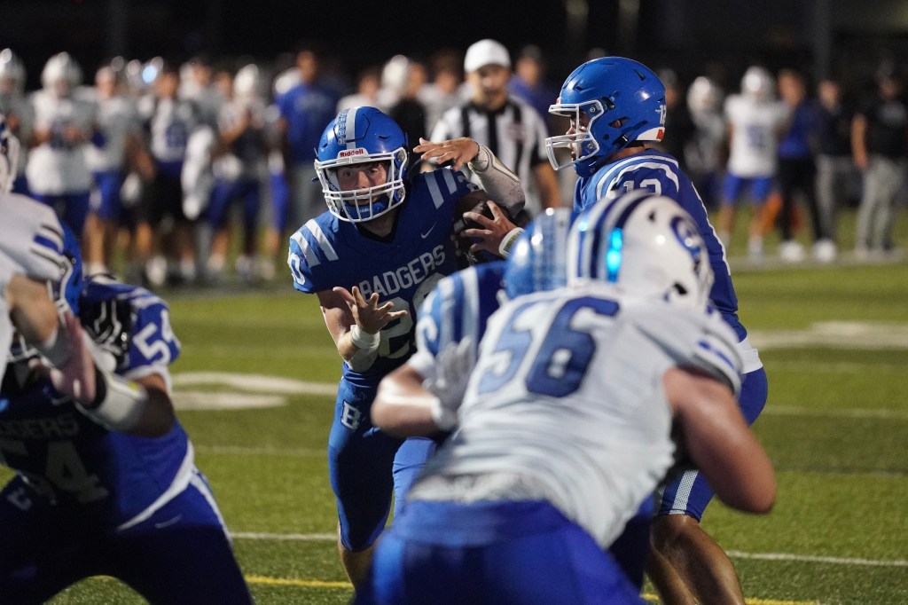 A high school football player in a blue uniform throws the ball while standing on the field. Opposing players in white uniforms attempt to tackle him in the foreground. A crowd watches in the background.
