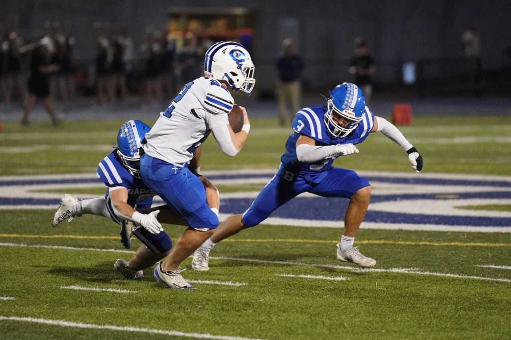 A football player in a white jersey dodges two defenders in blue jerseys during a game on a field.
