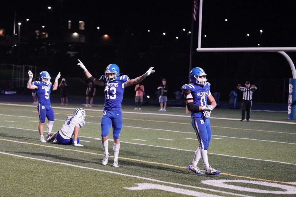 A group of football players in blue jerseys, celebrating on the field at night. One player with the number 33 raises both arms in victory, while another player stands beside him. A player in white is on the ground nearby.