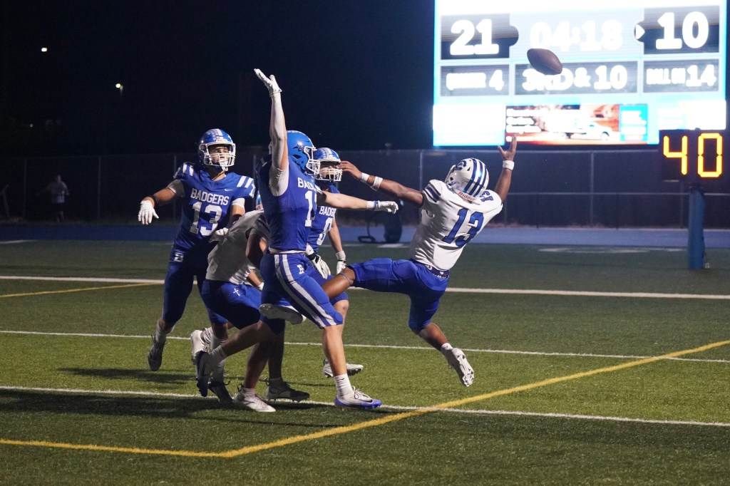 A football player in a blue jersey attempts to catch a pass while being defended by a player reaching out with his arm. The scoreboard in the background shows a score of 21 to 10 and indicates the game is in the fourth quarter.