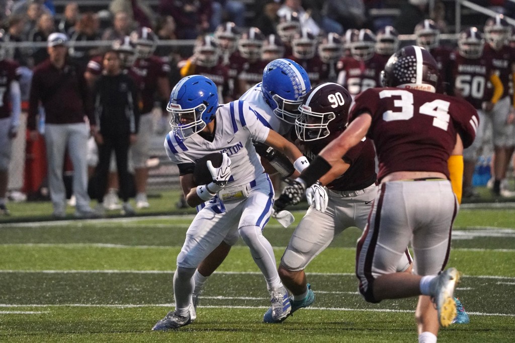 A high school football game in progress, featuring players in blue and white jerseys tackling an opponent in a maroon jersey on the field.