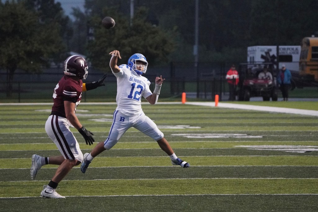 A football player in a white jersey with 'Bennington' printed on it is throwing a football while being pursued by a defender wearing a maroon jersey. The scene takes place on a football field in an evening setting.