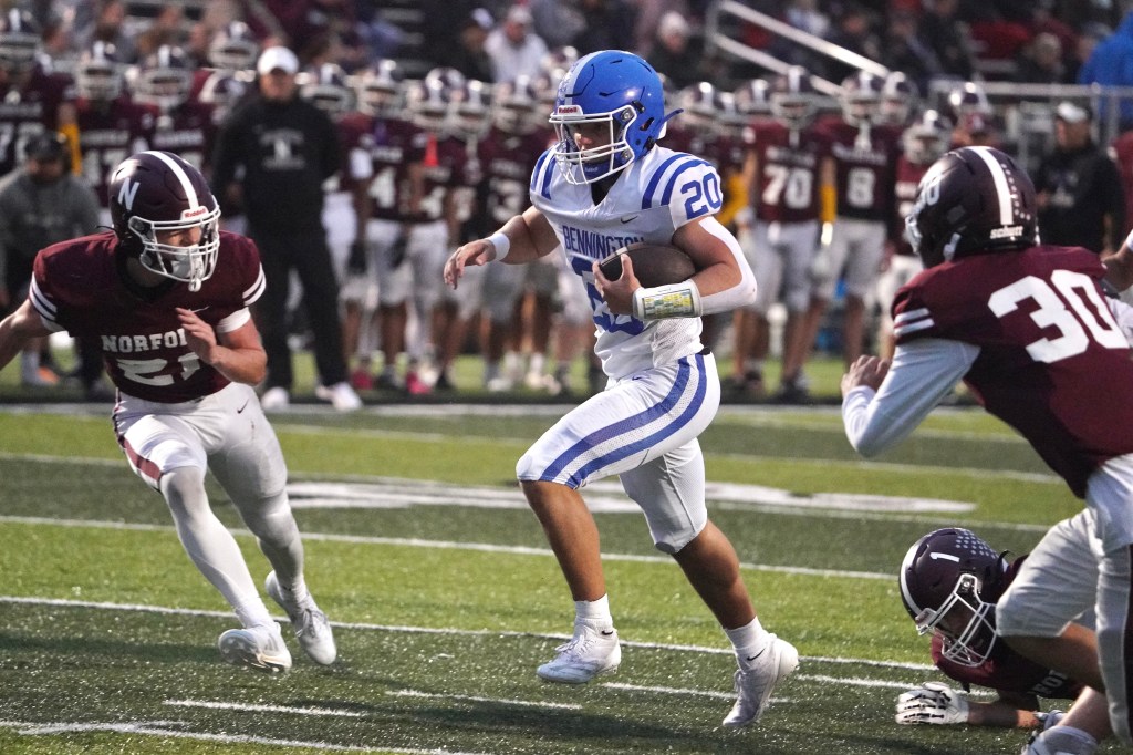 A high school football player in a blue uniform evades two defenders in a game, with a crowd of spectators in the background.