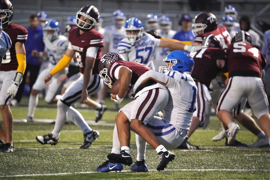 A high school football game in progress, showcasing a tackle where a player in maroon is being brought down by a player in blue. Other players are visible in the background, highlighting the competitive atmosphere.