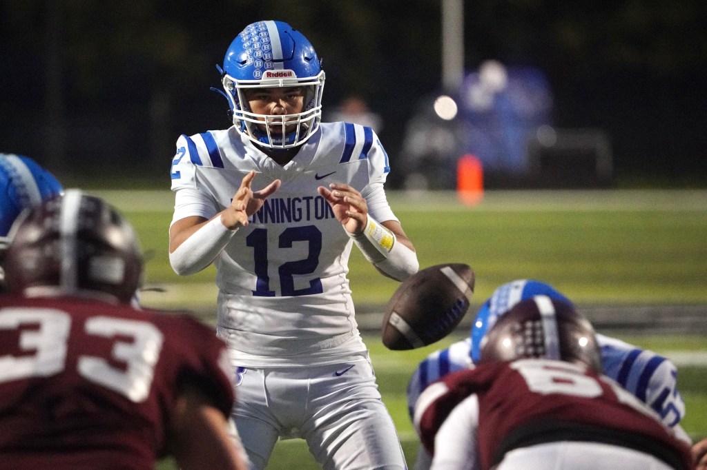 A football player in a white uniform with blue stripes, wearing a helmet, prepares to pass the ball on the field during a night game, with two opposing players crouching in front.