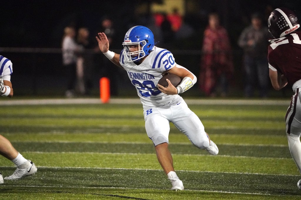 A football player in a white uniform with blue accents, wearing the number 20, runs with the ball while evading defenders on a sports field at night.