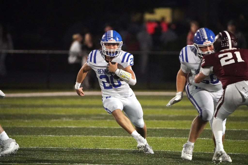 A high school football player in a white and blue jersey, number 20, runs with the ball while being pursued by defenders during a night game.
