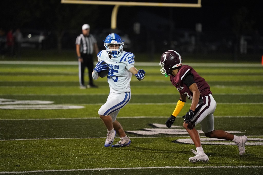A football player in a blue and white uniform evades an opponent in a maroon uniform during a night game on a well-lit field.