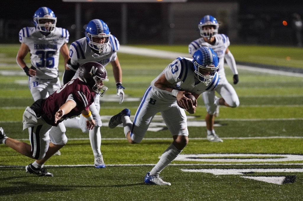 A football player in a white uniform evades a tackle while running with the ball, as other players in blue and maroon jerseys pursue him on a well-lit field.