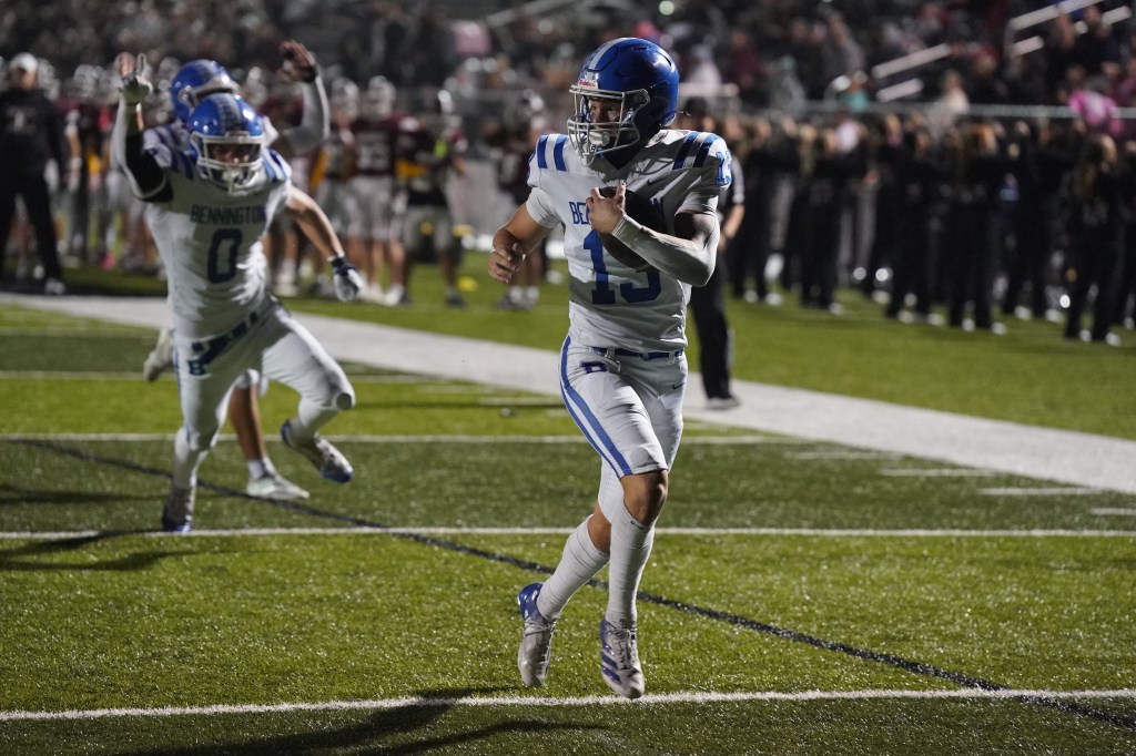 A football player wearing a blue and white jersey runs on the field during a night game, with teammates celebrating in the background.