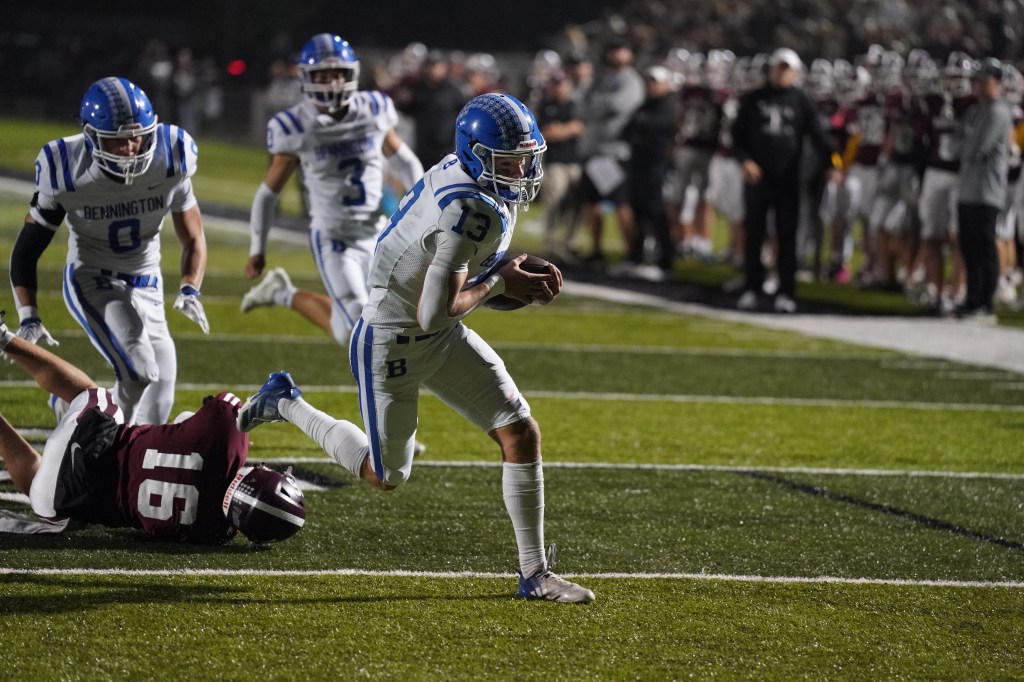 A football player wearing a white and blue uniform evades a tackle from a player in maroon while running with the ball during a game at night, with spectators in the background.