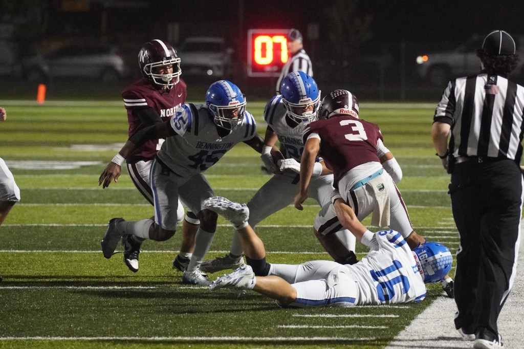 A nighttime high school football game scene, featuring players in blue and maroon uniforms. One player in blue is lying on the ground, while others are engaged in a tackle, with a referee observing the action in the background.