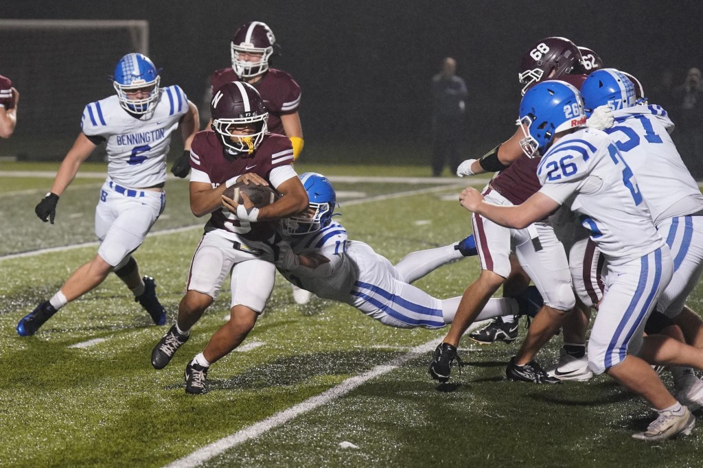 A high school football game in progress, with a player in a maroon jersey being tackled by defenders in blue and white jerseys on a wet field.