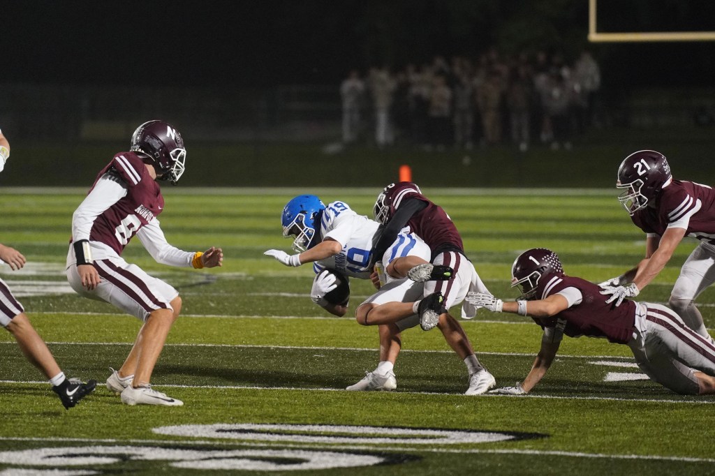 A high school football game scene with players in blue and maroon uniforms. A blue player is being tackled by two maroon players while another maroon player is in the background. The field is illuminated under the night sky.