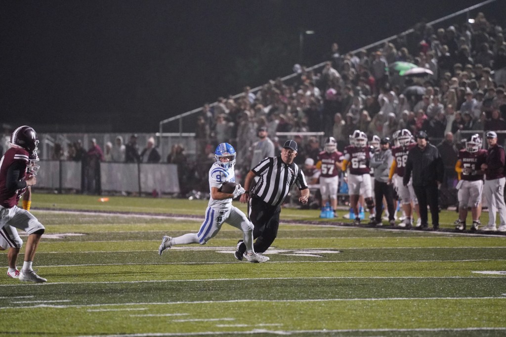 A high school football player in a blue jersey evades a referee while running on the field during a nighttime game. The crowd in the background is watching the action closely.