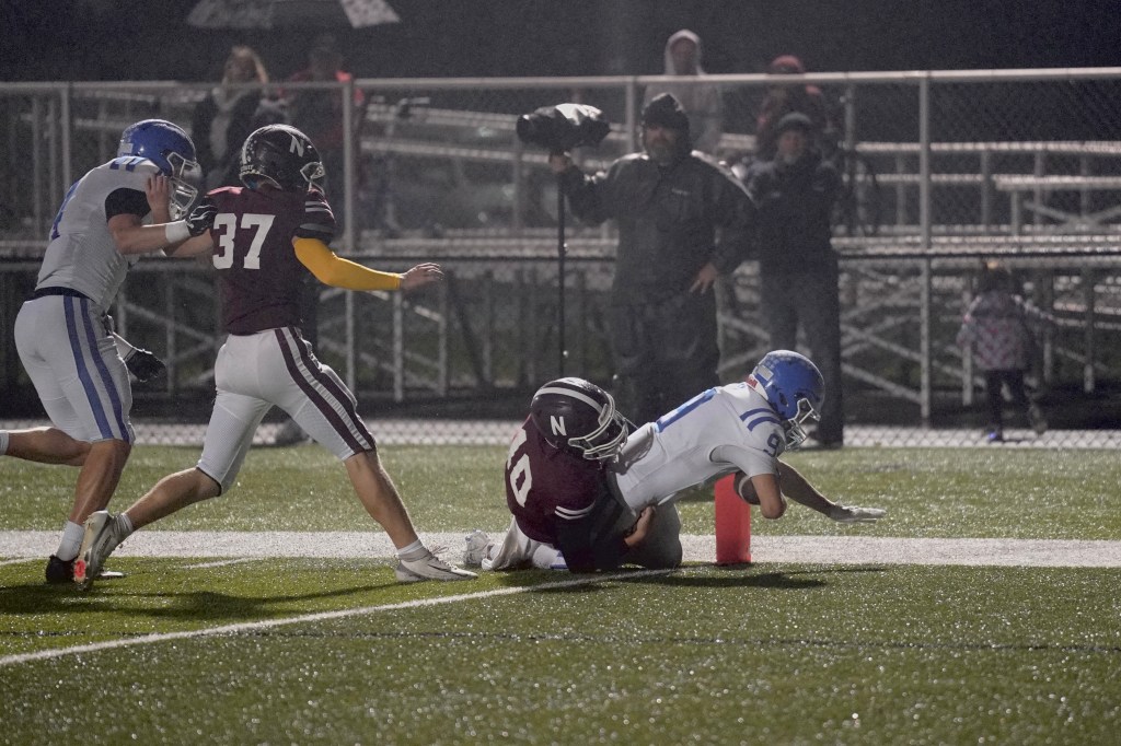 A high school football game scene with players in action. One player in a maroon jersey is diving toward the goal line while being tackled by defenders in blue jerseys. Spectators are visible in the background, and the field is illuminated at night.