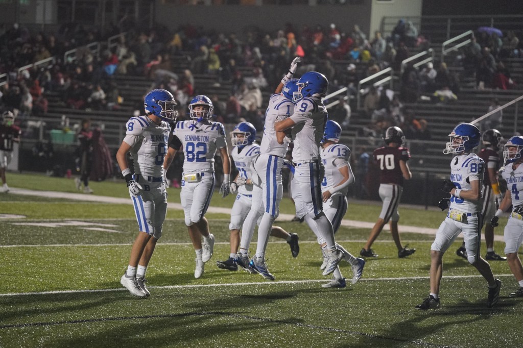 A group of high school football players in white and blue uniforms celebrating a touchdown on a night game, with spectators in the background.