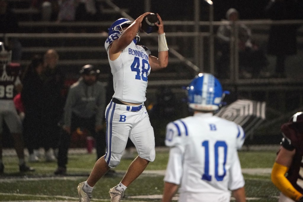 A high school football player wearing a white and blue jersey, number 48, prepares to throw a football during a night game. Other players and spectators are visible in the background.