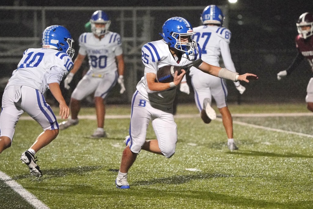A high school football player wearing a blue helmet and jersey runs with the ball, while teammates support from the left. The field is illuminated and wet from rain.