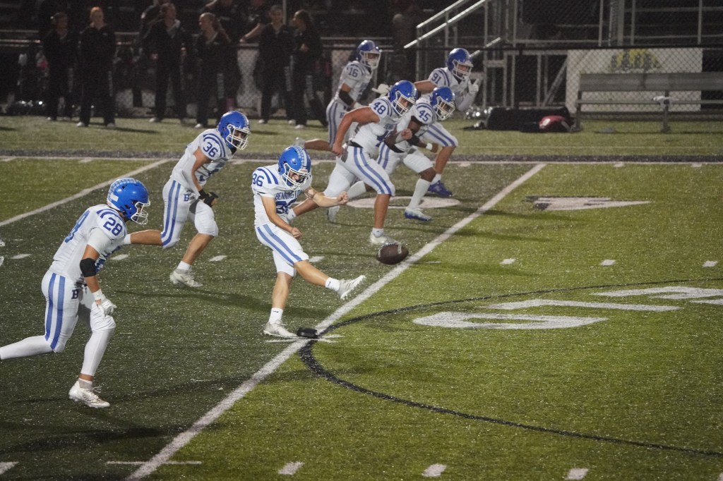 A football player wearing blue and white uniform prepares to kick the ball during a game, with teammates lined up behind him on the field at night.