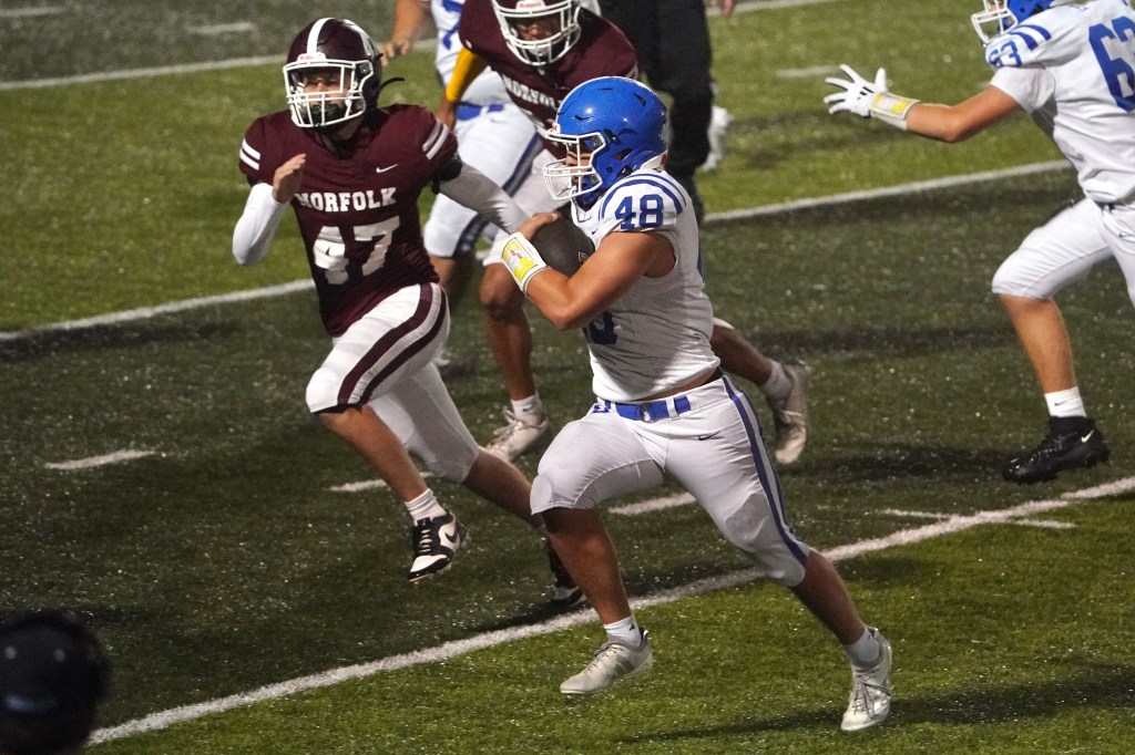 A high school football player in a blue jersey with the number 48 runs on the field while being pursued by a player in a maroon jersey with the number 47.