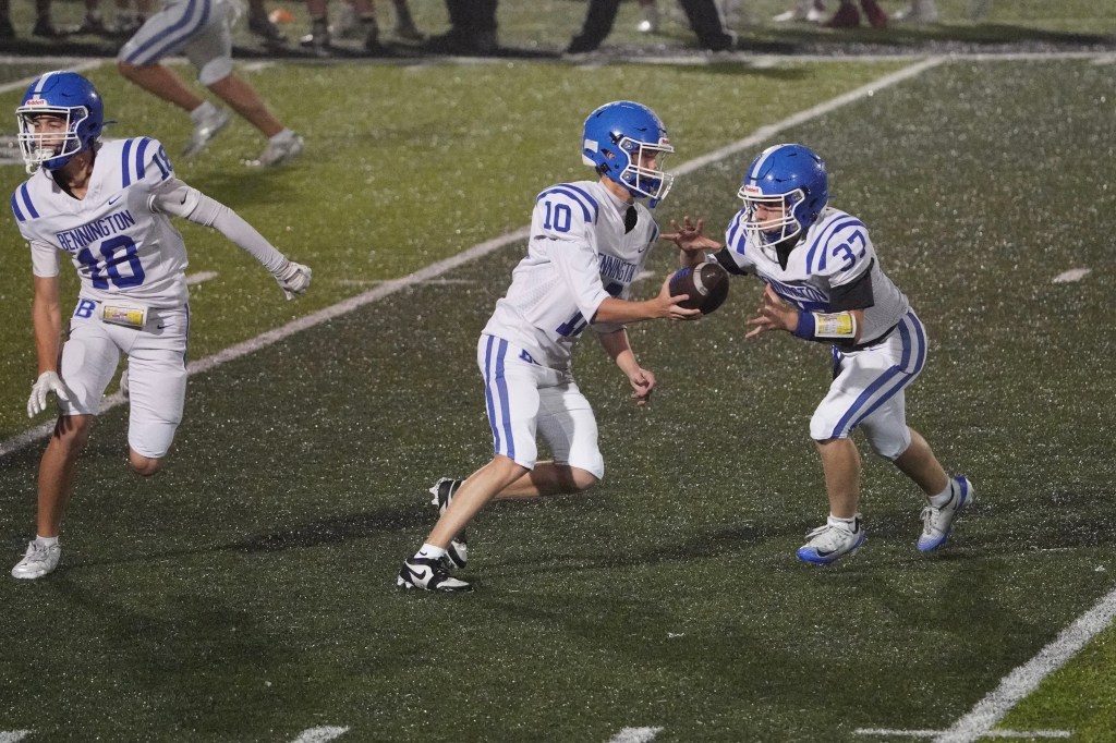 A high school football game in progress, featuring two players in white uniforms with blue accents exchanging the football, while another teammate stands nearby. The field appears wet, with a lively atmosphere.