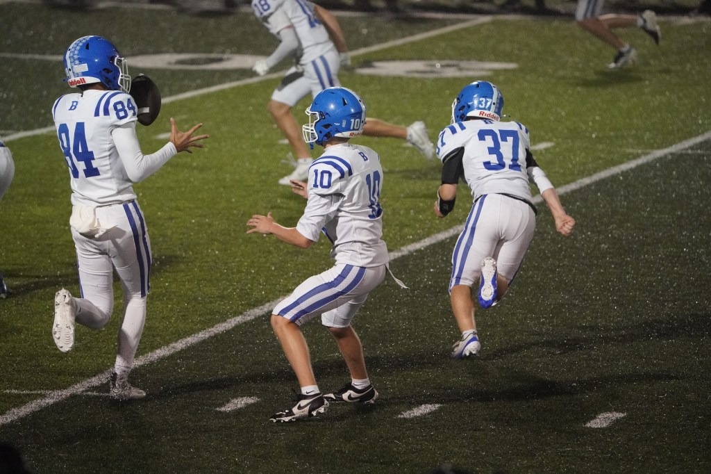 Three football players in blue and white uniforms running on a field during a night game.
