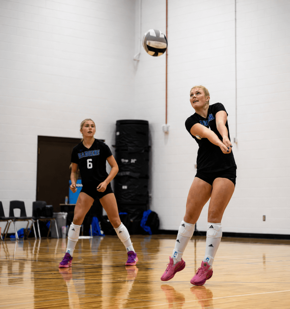 Two female volleyball players practicing in a gym. One player is preparing to receive a volleyball, while the other is watching. Both are wearing black t-shirts and athletic shorts, with knee pads and colorful shoes.