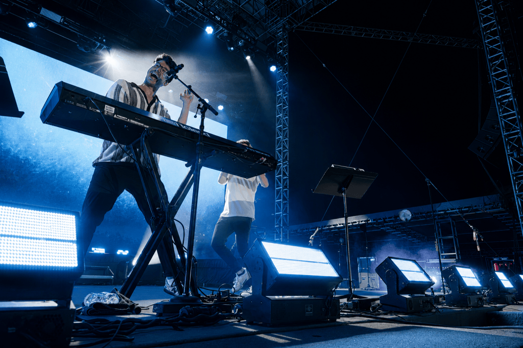 A musician performing on stage at a concert, playing a keyboard and singing into a microphone. The setup includes bright stage lights and a large backdrop with visual effects.