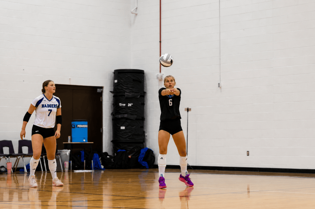 A volleyball player in a black uniform prepares to receive a serve, while another player in a white 'Badgers' jersey stands ready nearby in a gymnasium setting.