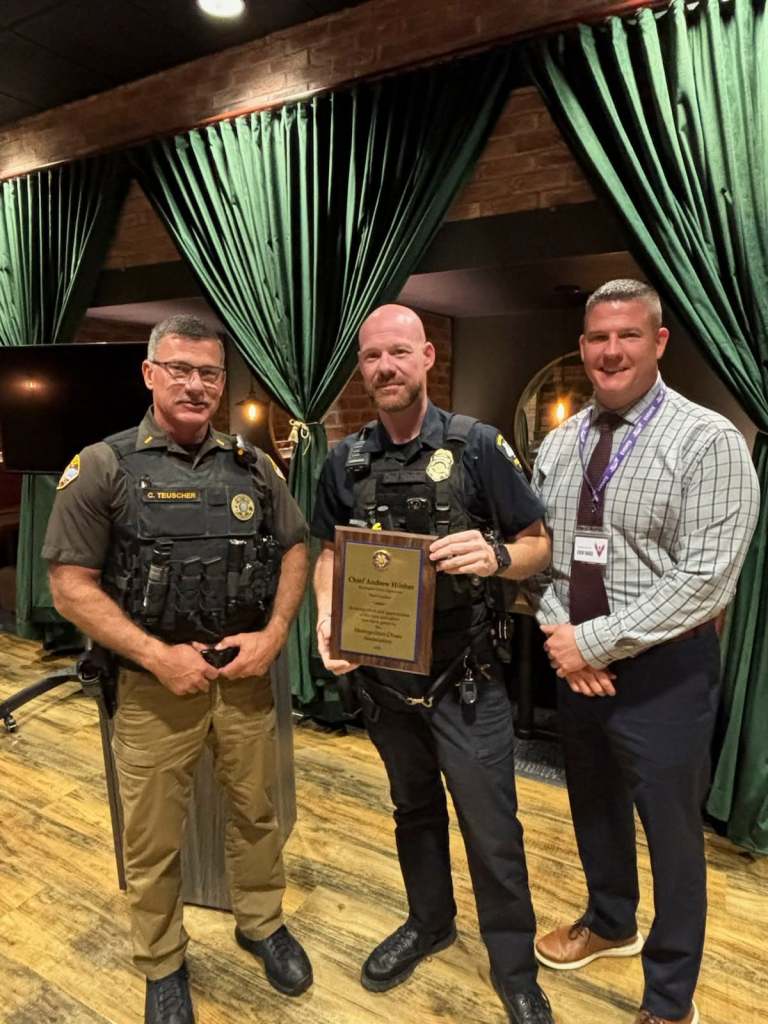Three men posing for a photo in a venue with green drapes. One man is holding a plaque, while the others stand beside him, smiling.