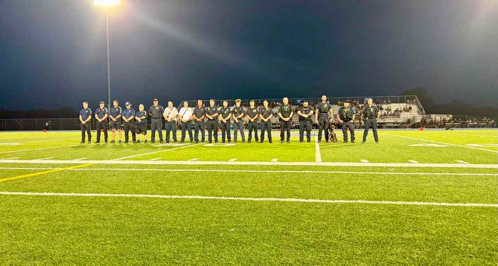 A group of uniformed personnel standing in a line on a football field under stadium lights during the evening.
