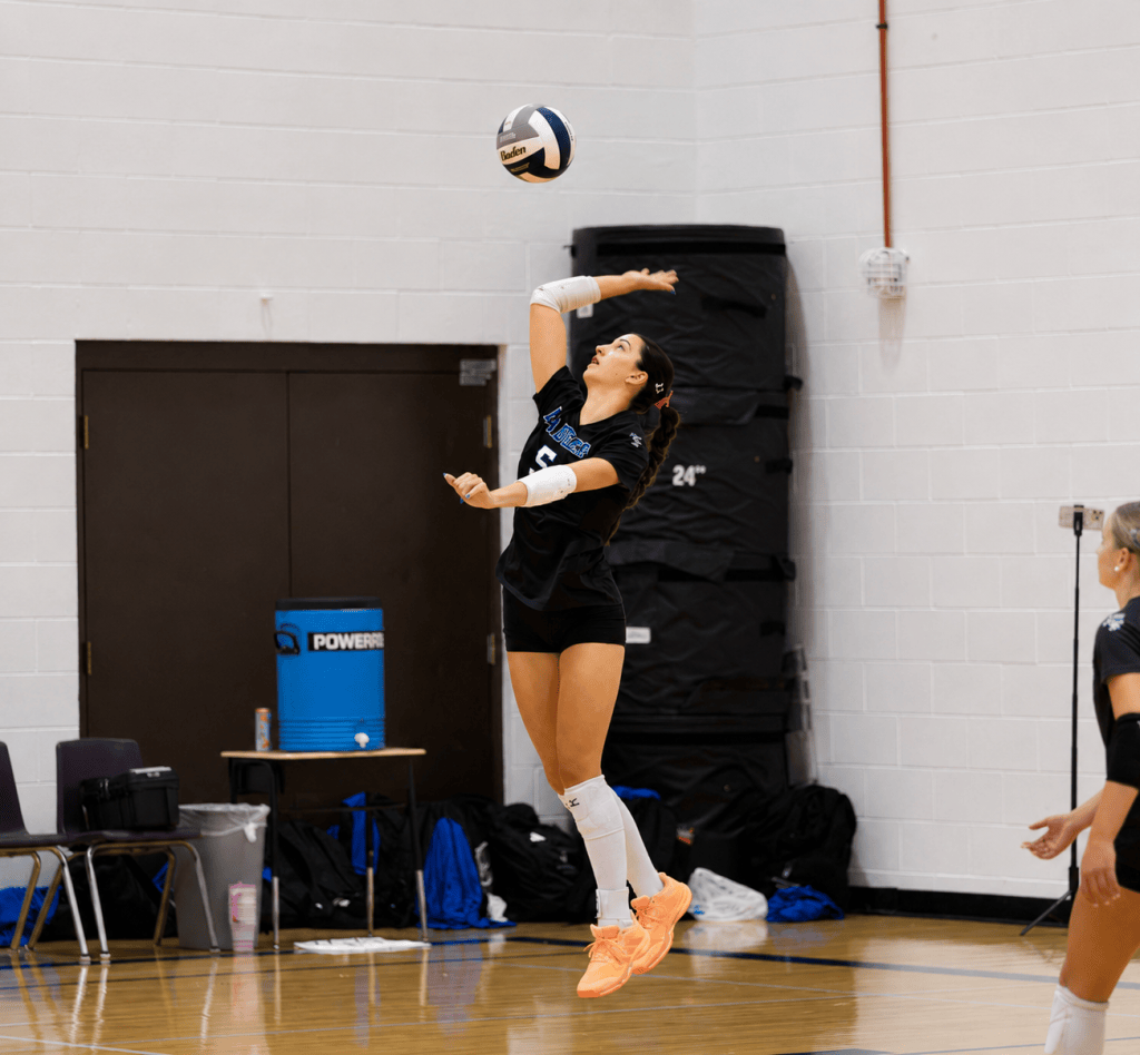 A volleyball player in a black uniform jumps to serve the ball, with a focused expression and arms raised. In the background, teammates are positioned ready for the play.
