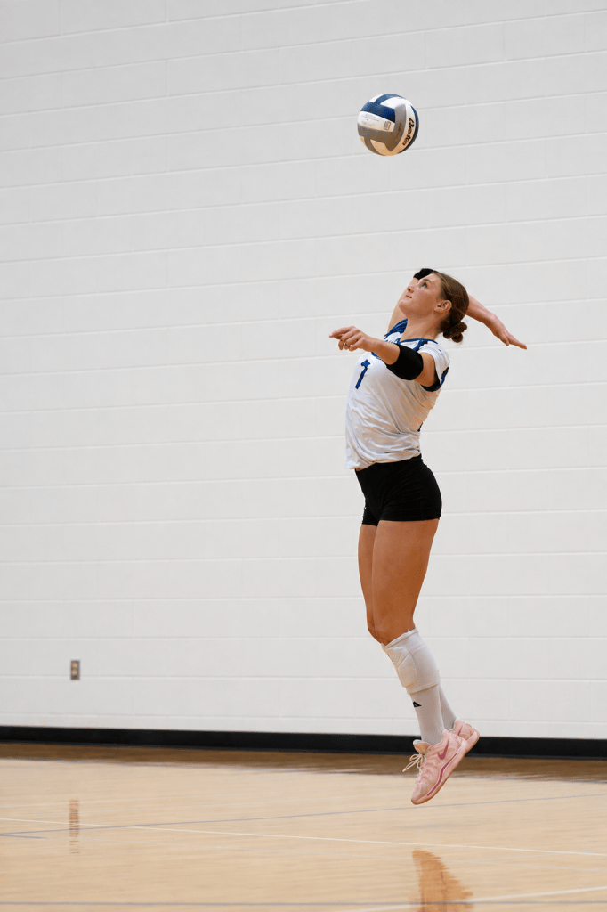 A female volleyball player jumping to serve a volleyball in a gymnasium setting.