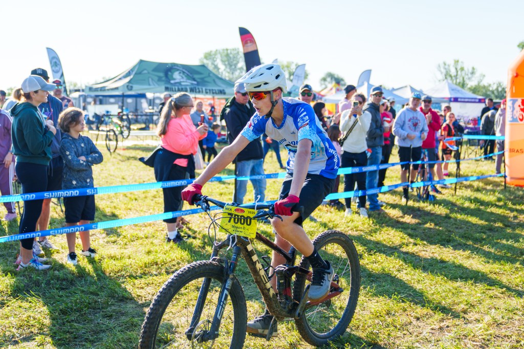 A young cyclist in blue attire rides a mountain bike through a grassy area, with a crowd of spectators watching in the background during an outdoor cycling event.