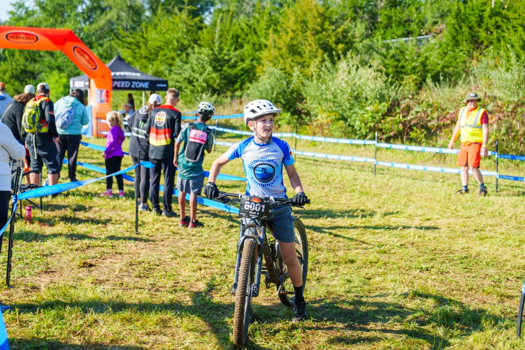 A young boy wearing a blue jersey and helmet smiles while standing beside his mountain bike at a cycling event. In the background, a line of spectators and participants is visible near a colorfully decorated archway.