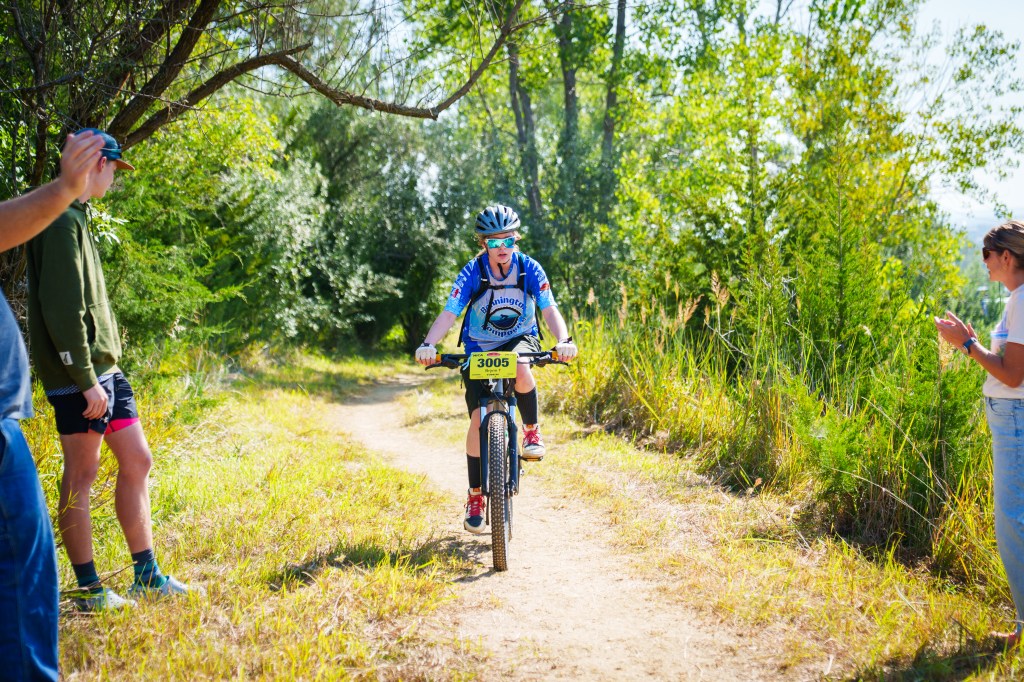 A cyclist wearing a blue jersey with the number 3005 rides on a dirt path surrounded by greenery, while spectators cheer and take photos.
