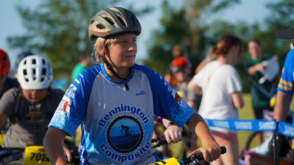 A young boy wearing a bicycle helmet and a blue cycling jersey stands next to a mountain bike during a race event, surrounded by other participants and spectators in a sunny outdoor setting.