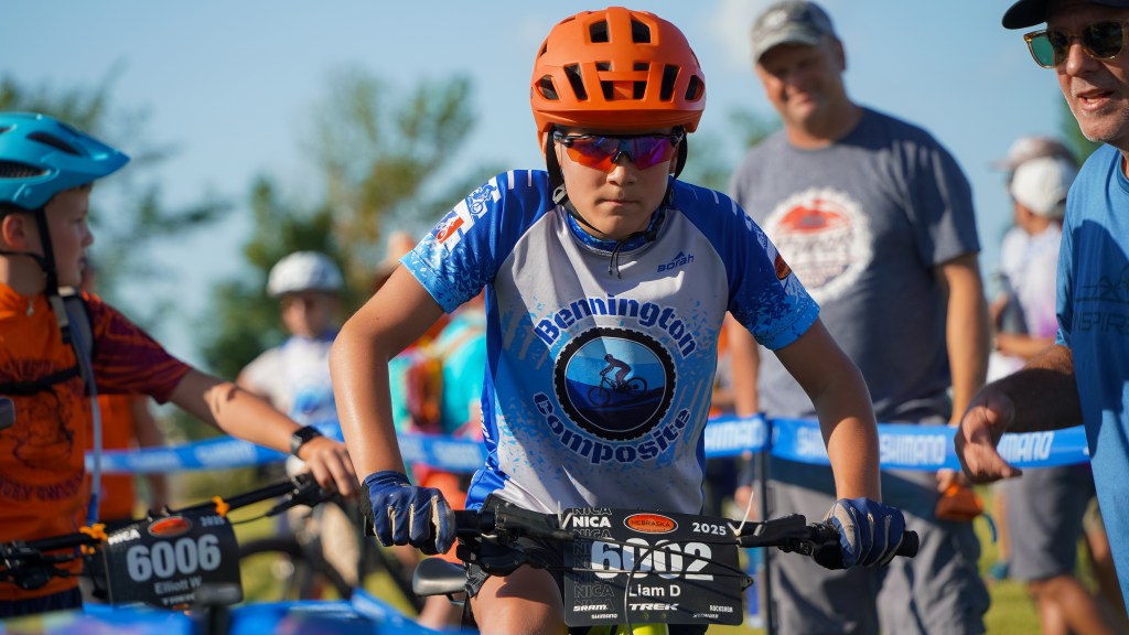 A young cyclist wearing an orange helmet and sunglasses prepares to race on a mountain bike, surrounded by other competitors and spectators at a cycling event.
