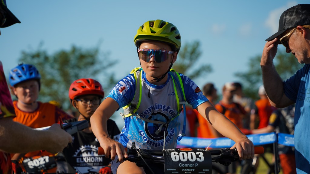 A young cyclist in colorful gear and a helmet prepares for a race, surrounded by other competitors and adults giving support.