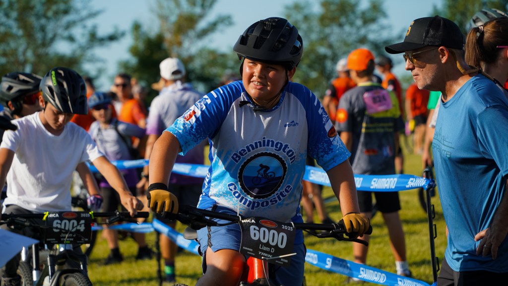 A young boy in a blue cycling jersey and helmet smiles while standing next to his bike at a cycling event. He's surrounded by other participants and spectators, with a vibrant outdoor background.